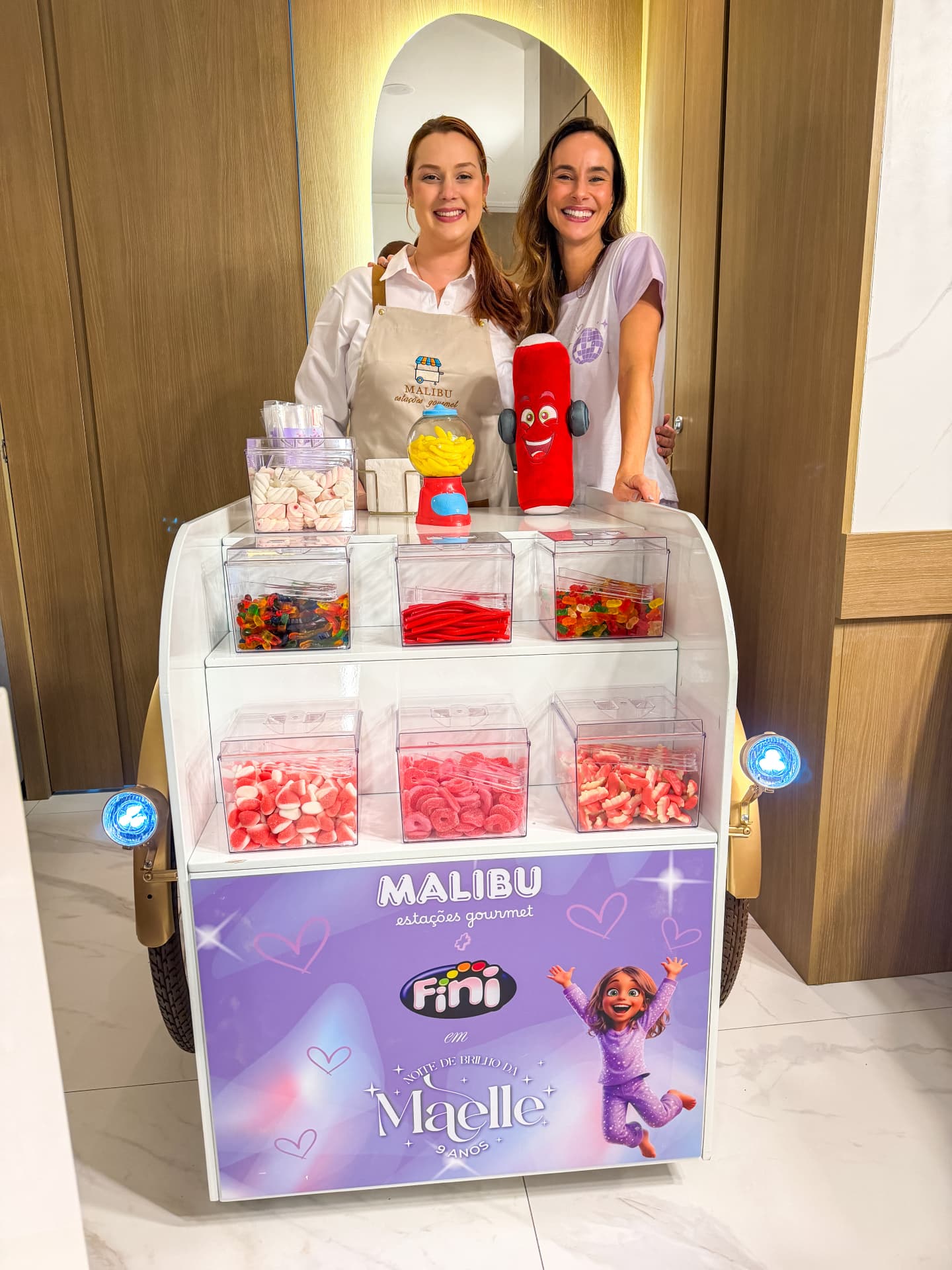 Equipe da Malibu Estações Gourmet ao lado do carrinho de doces personalizado em ação com a marca Fini