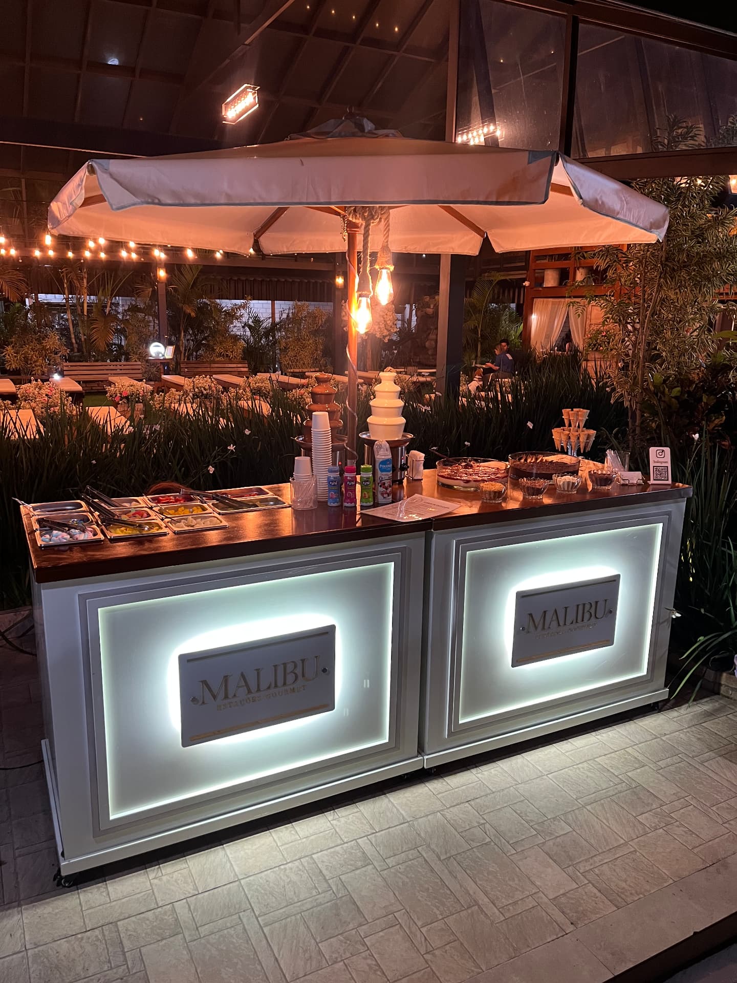 Estação gourmet iluminada em evento noturno, com fondue de chocolate, sorvete e coberturas
